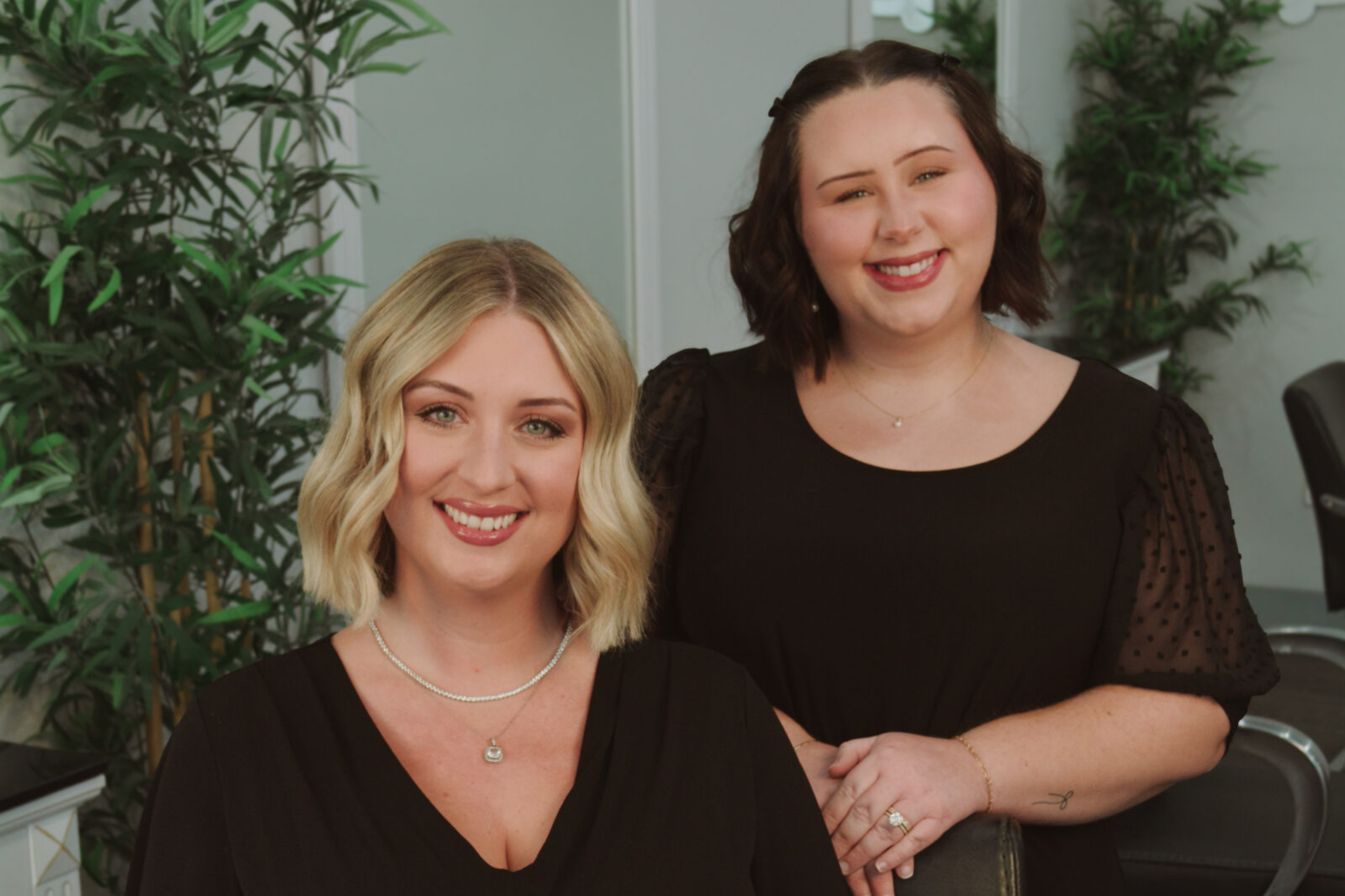 Two smiling women posing indoors with plants in the background.